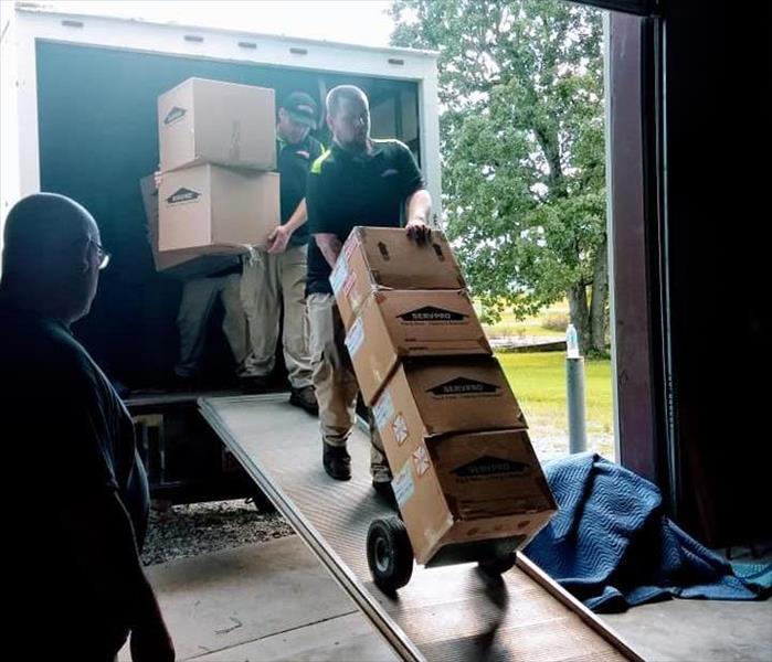 A man in SERVPRO uniform uses a dolly to bring boxes off of a moving truck while other men carry boxes behind him.