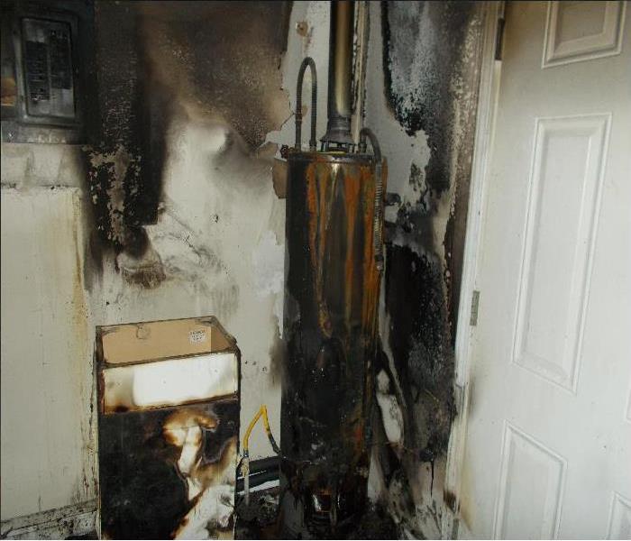 A burned up water heater sitting next to a white burned up cabinet. Soot covers the cabinet, walls, and water heater.