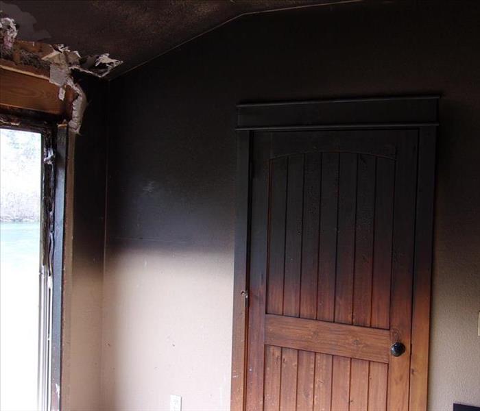 A cream colored wall and a brown door with heavy black soot halfway down the wall and door. Windows on the left are broken.