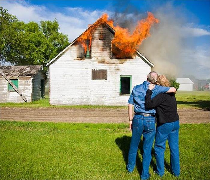 An adult man and woman stand holding each other while watching a white house burn.